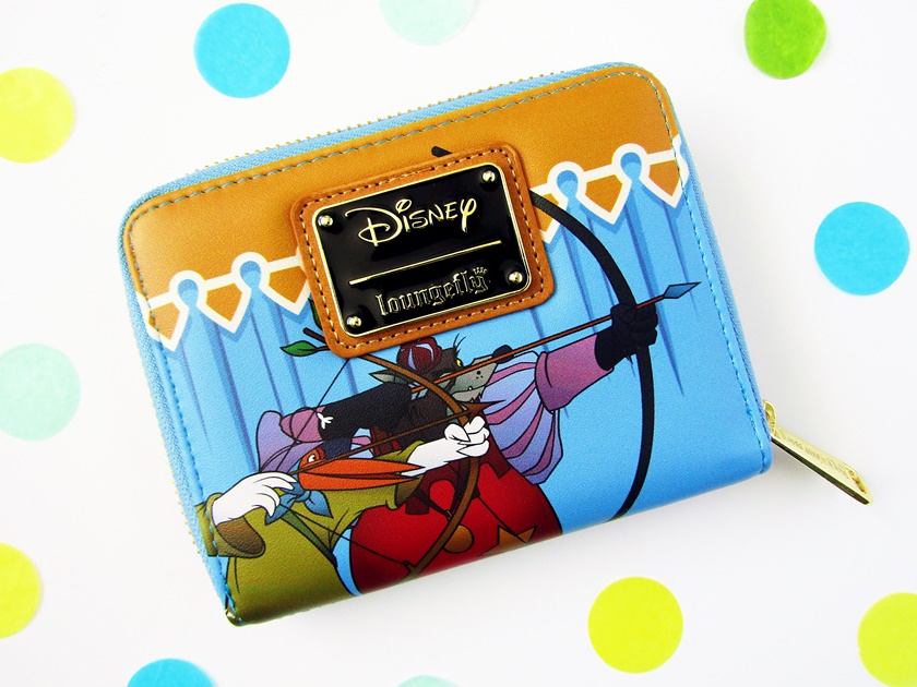 A photo of a blue and yellow Loungefly wallet with characters from Disney's Robin Hood printed on the front photographed from above on a white background surrounded by confetti. The front of the wallet features Robin Hood the fox dressed up as a stork and the Sheriff of Nottingham behind him, both holding bows and arrows as they take part in an archery tournament.