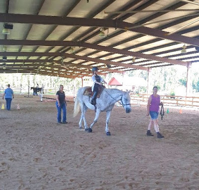 An image of a young woman, who is wearing a blue helmet and riding a white horse. They traverse a sandy floor, within a covered riding area, followed by a lady.