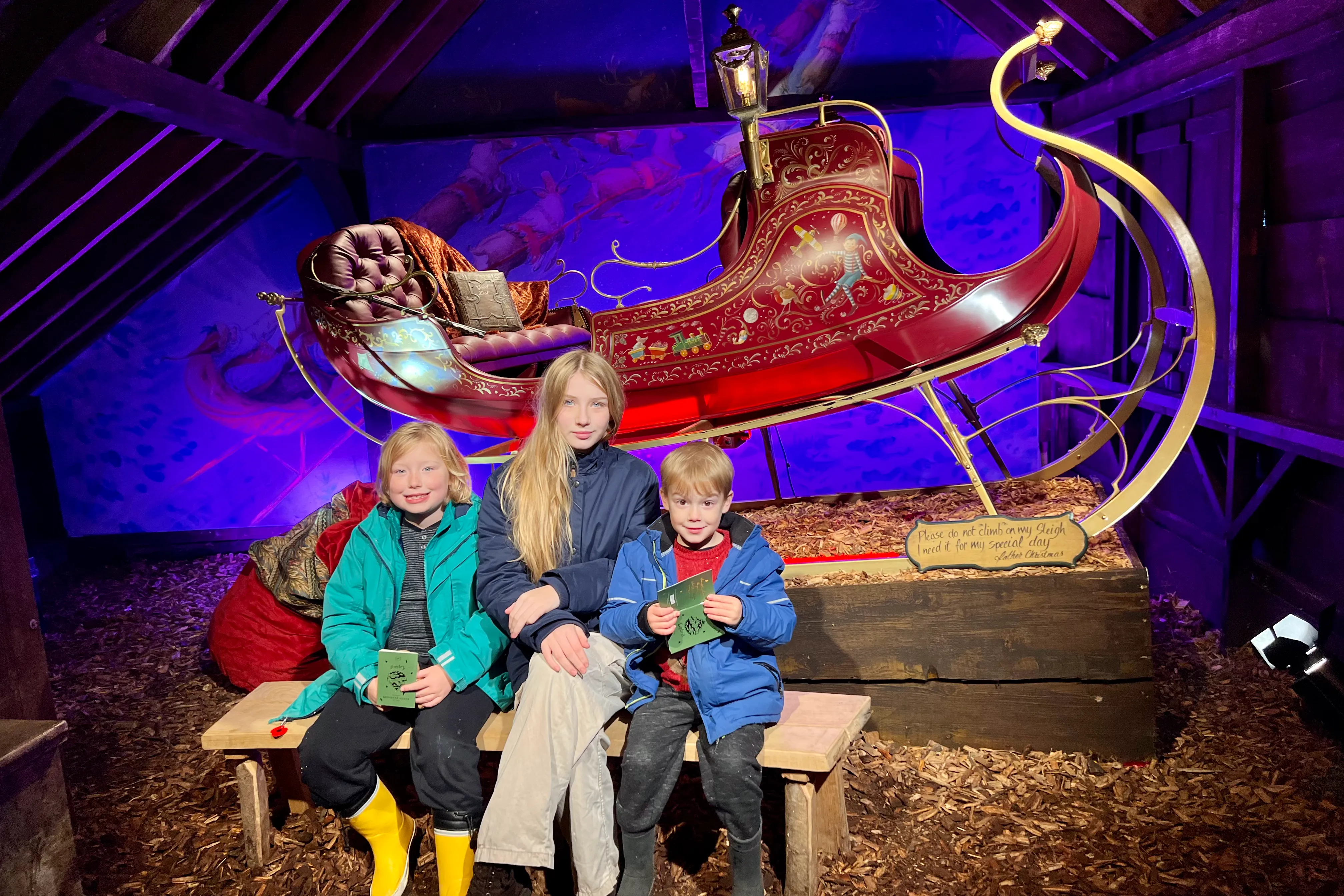 3 children sitting on a bench in front of Father Christmas' sleigh
