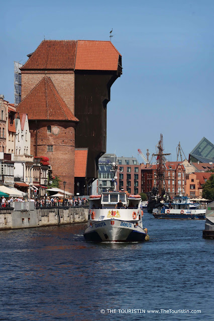 A white and black passenger ferry, and a dark wooden tall ship on a river lined by three-storeyed red brick period houses, a 15th-century red-roofed dark wooden crane structure, a red-roofed red brick stone tower, and a modern glass cube property, under a blue sky.