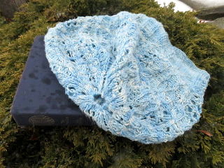 A fingering-weight cable and lace beanie hat laying flat. The hat has a lengthy ribbed rib that flows into intricate cables and lace.