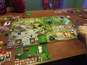 A game of Architects of the West Kingdom in progress. The board is laid out on the table, with player mats, cards, tokens, and meeples around the board and in various places on the board itself.