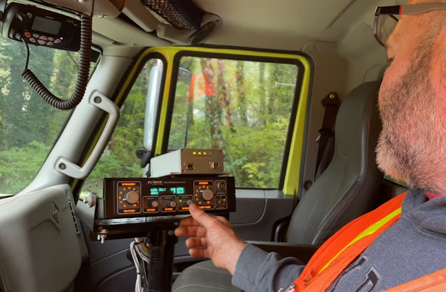 A person sits in the cab of a snowplow. They are reaching across the dash to operate a computer screen.