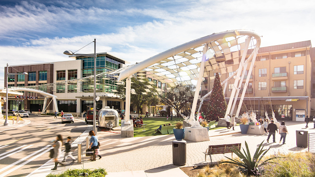 An expansive urban plaza within a modern land development project, illuminated by the sun. A distinctive architectural structure with white curved beams supports a geometric canopy, creating a dynamic play of light and shadow. The area bustles with activity, as people walk along the pedestrian crosswalks and relax on avant-garde benches. The backdrop features a blend of contemporary and traditional architecture, including a storefront with the "Subway" sign, indicating a mix of retail and residential spaces. A large, decorated Christmas tree suggests a festive season, adding warmth to the community-centric design. The scene encapsulates a successful transformation of space through thoughtful land development, emphasizing usability, aesthetics, and social interaction.