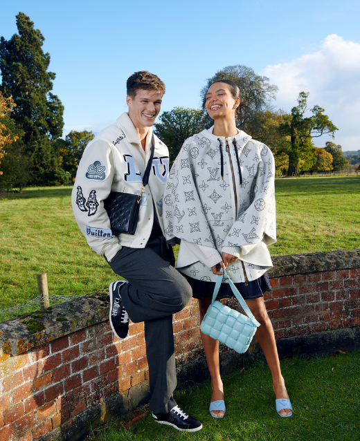 Male and Female model in outdoor grassy scene laughing. Both models are wearing designer outfits including a Louis Vuitton varsity jacket, Dior trainers and a Bottega handbag