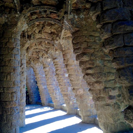 A view under and between ancient stone columns. The columns on the left are vertical, and the roof arches over tothose on the right that lean out at a 30 degree angle. Sunlight is coming through the stone columns on the right, the shadows falling across the ground in a striped fashion. The location is the  in Park Güell, a stunning park in Barcelona that was designed by Antoni Gaudí.