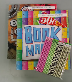 Two colorfully-decorated portfolios, one on top of the other and each printed with the words, 'Book Marks.' Strips of label-maker tape have been put across the top of each one, reading, 'COST: ONE ROCK-STAR EACH.' Sitting on top of the two portfolios are several novelty-printed pencils, held together in clear-plastic packaging. From left to right, the pencils are pink-patterned, light green-patterned, black-against-pale-green zebra-patterned, and money-patterned, with about four of each variety visible.