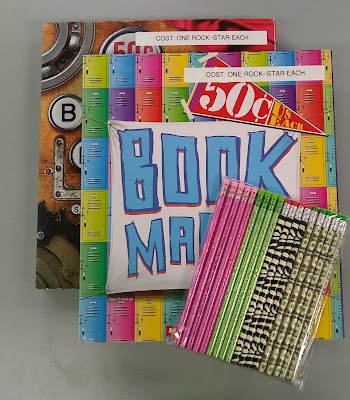 Two colorfully-decorated portfolios, one on top of the other and each printed with the words, 'Book Marks.' Strips of label-maker tape have been put across the top of each one, reading, 'COST: ONE ROCK-STAR EACH.' Sitting on top of the two portfolios are several novelty-printed pencils, held together in clear-plastic packaging. From left to right, the pencils are pink-patterned, light green-patterned, black-against-pale-green zebra-patterned, and money-patterned, with about four of each variety visible.