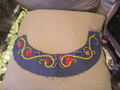 A half-circle band of blue linen with a mirrored vining design in yellow beads with blue leaves and red, white, and black flowers, still fairly sparse, laid on a tan couch pillow.