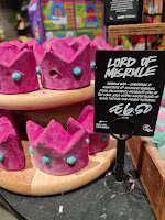 A photo of a square light brown box filled with a bunch of red and gold crown shaped bubble bars next to a rectangular black card that says Lord of Misrule bubble bar in white font on a light brown rectangular table  on a bright background