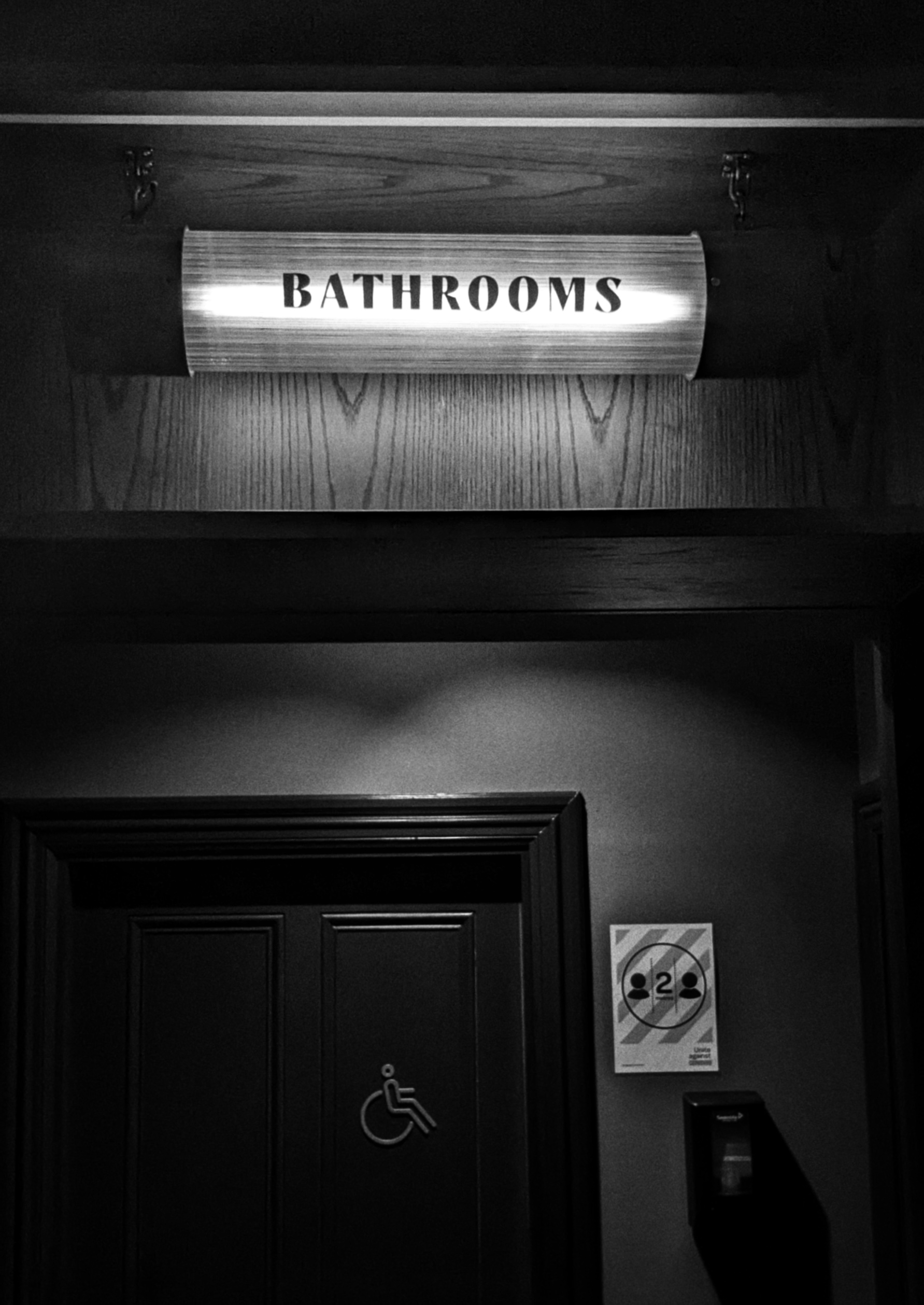 Black and white photo of a dark toilet door and 'Bathrooms' sign above