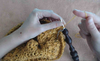 Someone crocheting around the edge of a crochet hat.  The yarn is yellow fingering-weight yarn with flecks of black, and is held with two strands together.  The crochet hook is metal with a wooden handle. 