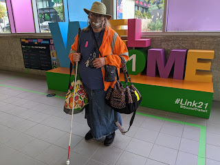 Tall white woman in orange jacket with tan hat and olive mask, various bags, white cane