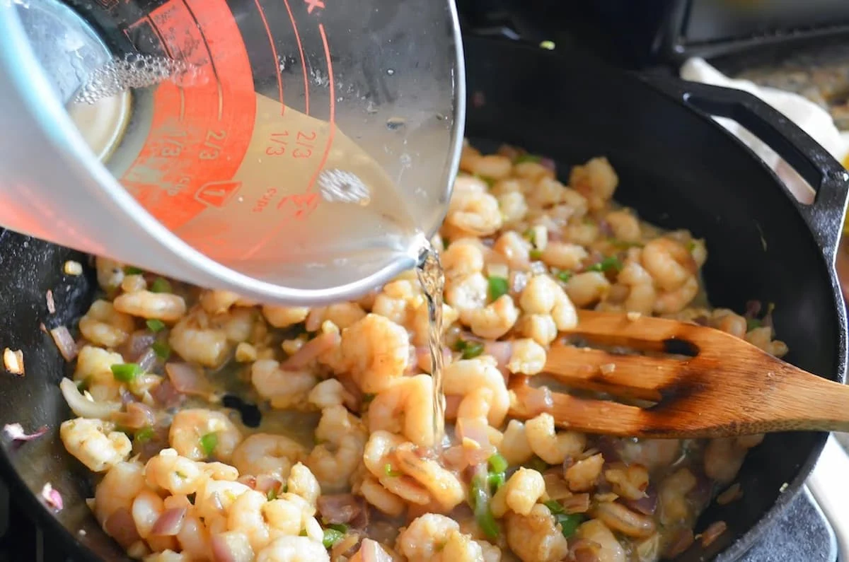 White wine being poured into shrimp mixture in a cast iron pan.
