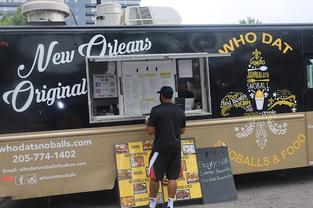 A man is ordering from the black and tan Who Dat Snoballs & Food truck. The side of the truck says "New Orleans Original."