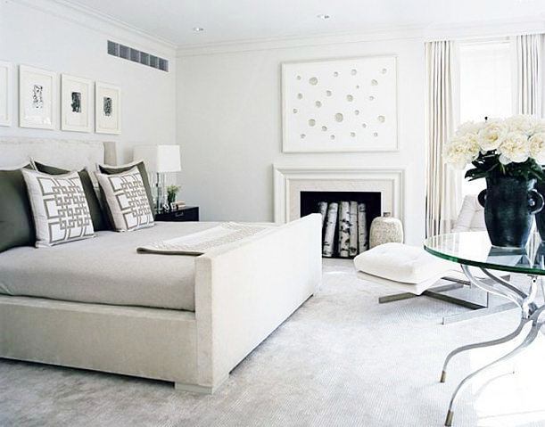 gray bedroom with upholstered bed, a glass table with a large jug of white roses, and molded white fireplace