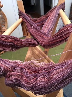 A folding chair standing upside down, with the legs half-folded, and pink, purple, and white yarn wrapped aorund the legs in an ex. The yarn drapes softly between the legs of the chair, and ripples slightly at points from the twist of the yarn now that it's loose. It looks squashy and nice to touch.