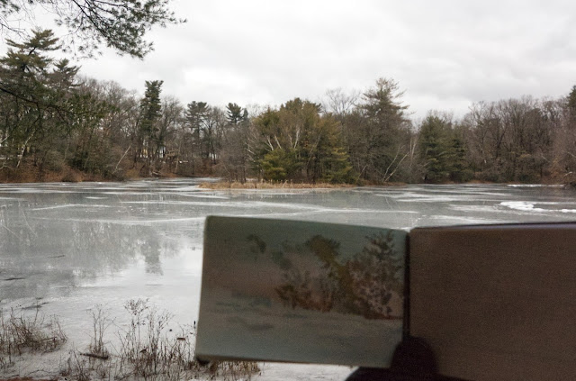 alt="Photo of wooded peninsula with icy pond and melted puddles in foreground and sketchbook with partially completed painting held up in front."