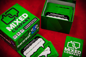 The game box, a green box just large enough to hold a couple hundred normal-sized playing cards. The box is green, with the name and logo on the cover. The cover sits next to the bottom, which holds a number of cards. Some of these cards are lying on the table next to the box. Some cards are face down, so you see the back, which consists of the name and logo. Others are face up, so you can see that they have a white speech balloon on a green background, with a variety of different text in the balloons.