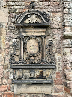 An ornate looking memorial on the wall of Duddingston Kirkyard.  On it are various emblems such as the skull and crossbones and the face with wings at the side of it.  Photo by Kevin Nosferatu for the Skulferatu Project.