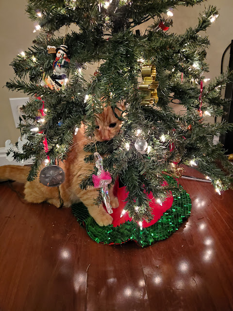 Orange cat chewing on a small, lit artificial christmas tree with ornaments