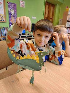 Uśmiechnięty chłopiec, pokazuje do zdjęcia wyko wykonaną przez siebie pracę plastyczną papierowego dinozaura. Tło sala biblioteczna i regały z książkami.