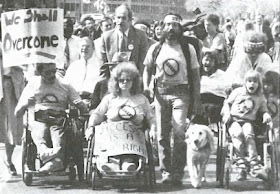 a black and white picture shows a crowd of people, some of whom use wheelchairs, one man has a service dog with him. They are children and adults and appear to be chanting or singing. One person holds a sign that says, “We shall overcome.”