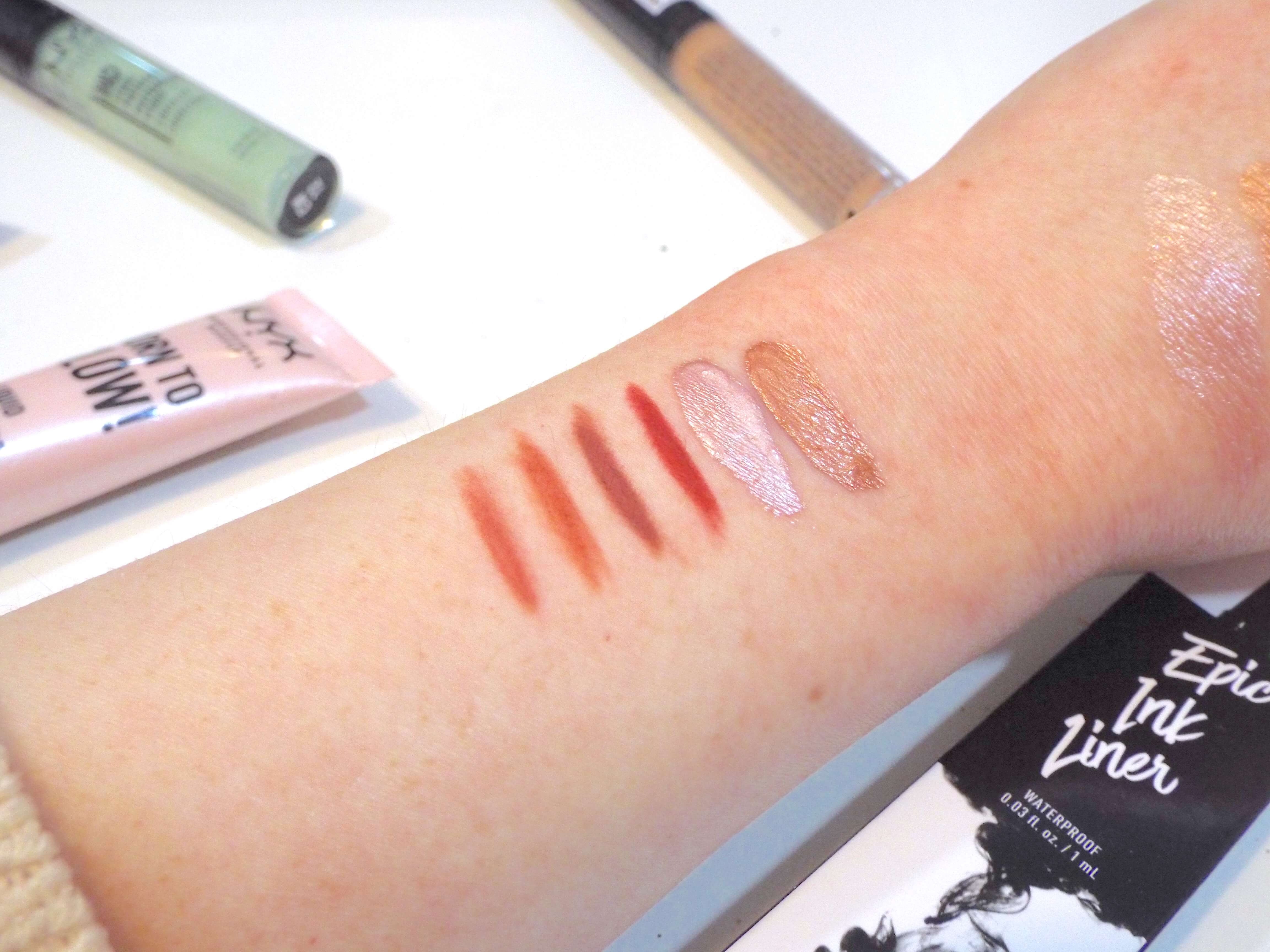 Swatches, left to right, of four NYX Lip Liners in a warm, pinky nude, warm orange nude, mauve-y pink and rose mauve-y pink, and two NYX Born to Glow Liquid Illuminators, in an icy pink shade then a deep warm golden shade.