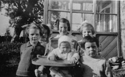Black and white photo of five girls and a baby outside a stone built house with a half glazed porch.  There is a large tree in the left background.  The baby is sitting in a highchair surrounded by the other girls