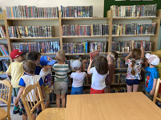Na zdjęciu grupa dzieci oglądająca książki, dzieci ustawione są tyłem do fotografującego, w tle znajdują się regały z książkami.