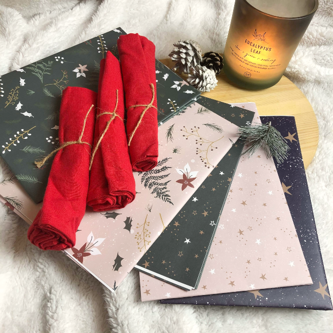 Four red napkins rolled up on top of multicolour Christmas wrapping paper next to a lit candle