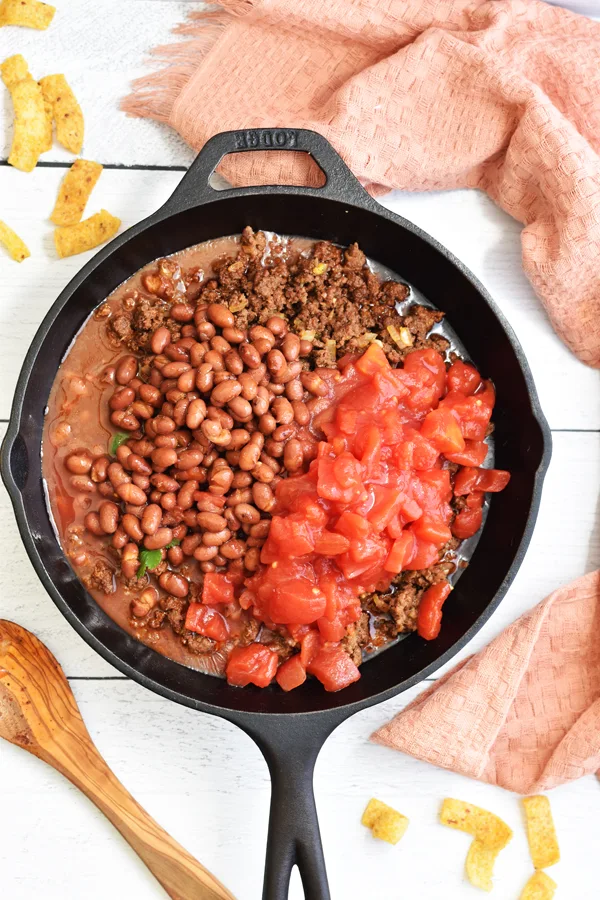 Diced tomatoes being added to browned ground beef in a cast iron skillet, enhancing the flavors and creating a delicious base.