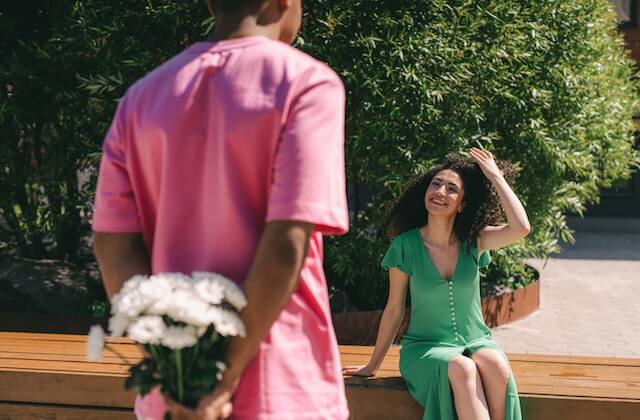 Woman in green dress sitting on the bench smiling to a man holding flowers in his back