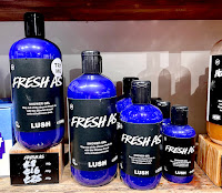 Selection of tall, cylindrical plastic play bottles filled with turquoise shower gel in varying sizes next to a light, brown and black square wooden block with Lush fresh as shower gel in white font. All gels have a rectangular black label with lush fresh as shower gel in white font on a bright background