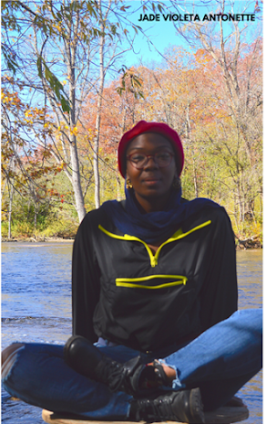 Jade Violeta Antonette, a dark-skinned Black person, sits on top of a wooden staircase outdoors with a shadow over her face. She is wearing a black top, blue jeans, and a red beret. There are trees with changing leaves in the background.