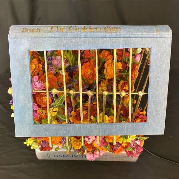 paper sculpture of The Golden Cage hardback book with cover cut out to show profusion of colorful paper flowers inside behind golden paper bars