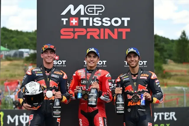 Podio del Tissot Sprint de MotoGP en Brno 2025: Marc Márquez (centro) celebrando la victoria junto a Pedro Acosta y Enea Bastianini (ambos de KTM) con botellas de Prosecco.