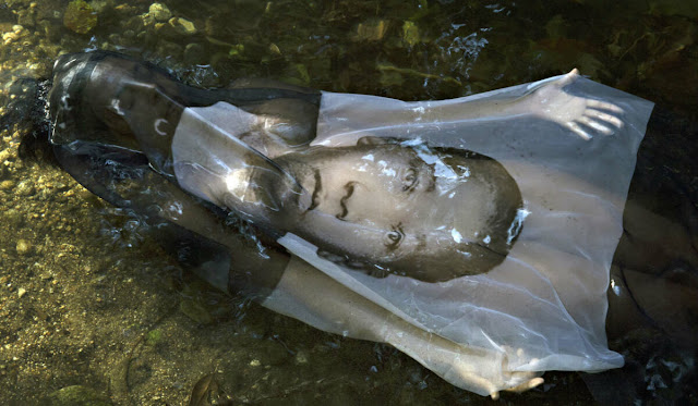 Mulher boiando na água, aparentemente de um rio, com um pano em cima de seu corpo  no qual está impresso o rosto de um menino negro. Este tecido é translúcido e por baixo dele é possível ver um pouco do corpo da mulher.