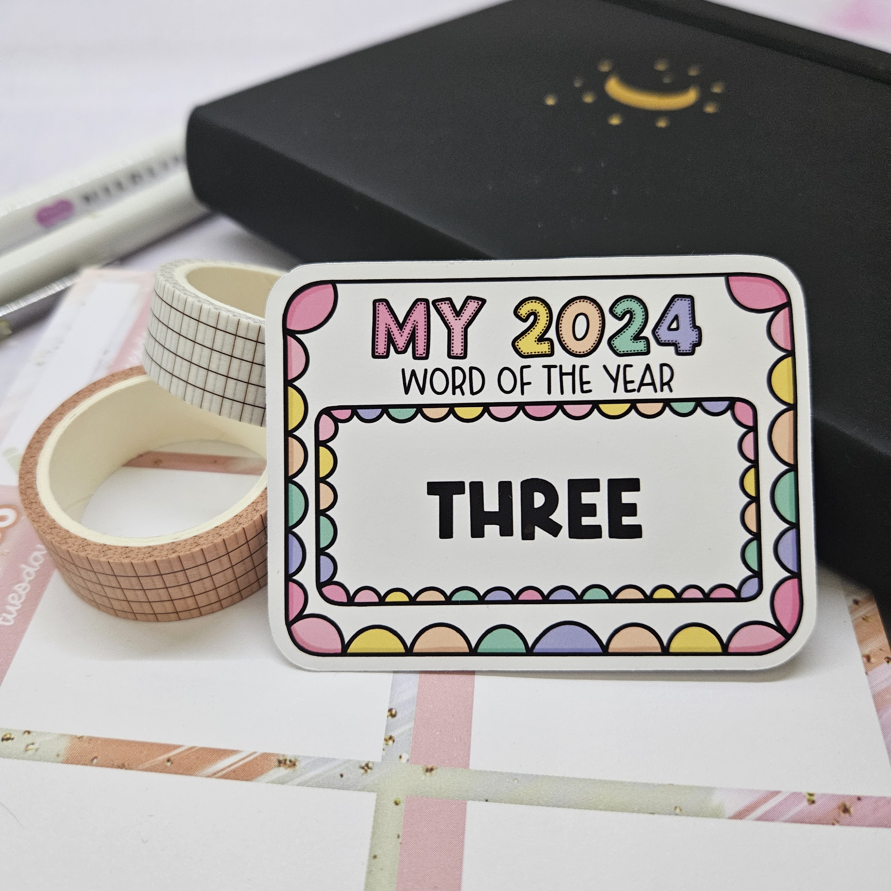 Image shows a sticker of the word of the year "three" with a black bullet journal underneath it and a notepad. There are also two pens and two washi tapes in the photo.