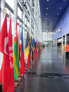 A photo looking down a long hallway with a glass wall to the left and a blue wall with a large entrance door to the right. In front of the glass wall is a long row of red, green, blue, and yellow flags hanging from short flagpoles resting on the floor. 