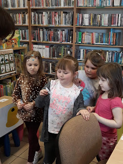 Grupa dziewczynek stoi , jedna z nich trzyma w dłoni obrazek. Tło: sala biblioteczna.