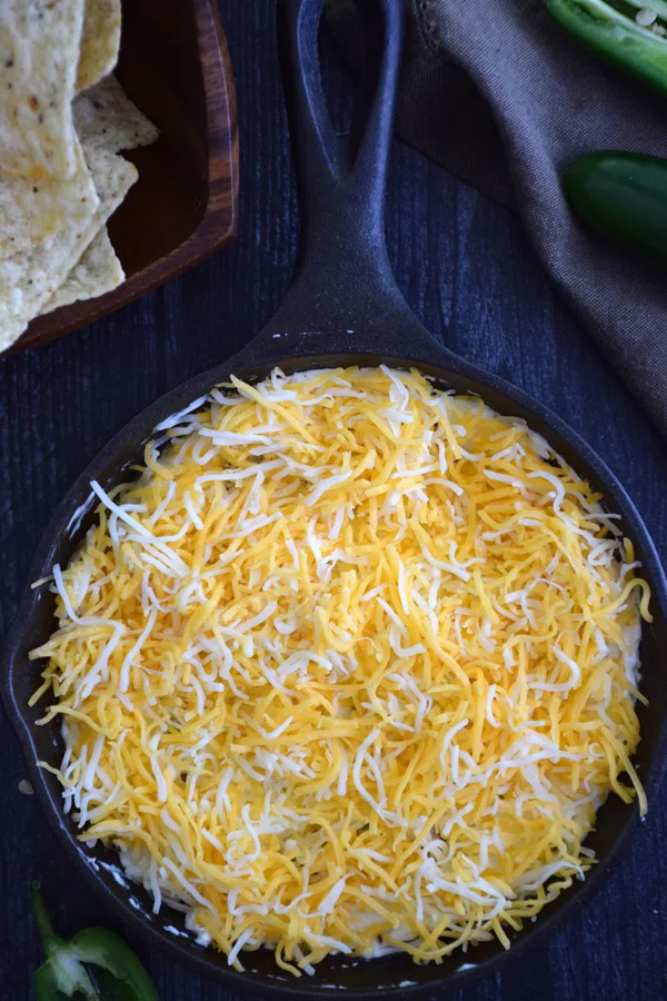 A tantalizing view of a cast iron skillet filled with jalapeno popper dip, generously topped with shredded cheddar cheese.