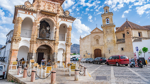Pequeña capilla y la plaza con la iglesia de Santa María, con cielo azul con nubes de fondo.