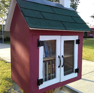 Cabinet of books, mounted outdoors on a post and designed to look like a little building. The cabinet has red siding and a roof covered with green shingling. Through glass doors that are framed in white-painted wood, two shelves of books are visible.