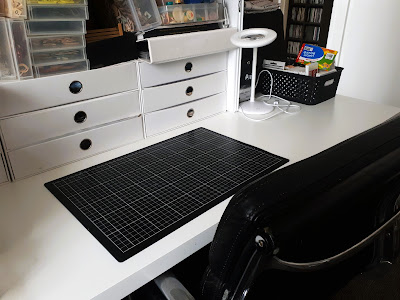 Clear work desk with a cutting mat on it, and a bank of storage drawers to the back of it with storage containers on top of them.