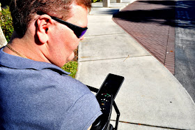 A power wheelchair wearing sunglasses looks down at his phone, which is mounted to a bracket on the right side, close to the wheelchair's joystick.