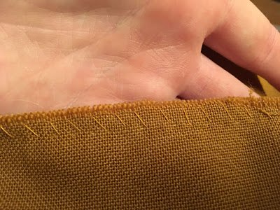 A close-up of a seam edge in mustard-brown fabric with a large, frayed allowance and huge whipstitches in bright yellow thread, supported by a white hand.