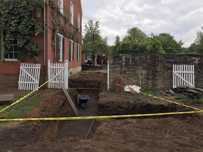 Multi-story brick building in upper right corner of photo. In forefront, several trenches have been dug for installation of pipes. Yellow caution tape blocks the trenches.