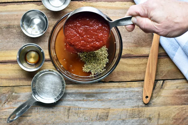 Various ingredients being mixed together in a glass bowl to make BBQ sauce, placed on a rustic wooden table. Ingredients include ketchup, chili sauce, brown sugar, vinegar, and spices, ready to be stirred into a sweet and tangy sauce.