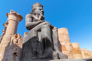 foto de uma estátua colossal de Ramsés II sentado, em cor cinza, com altura de 15,5 metros na entrada do templo